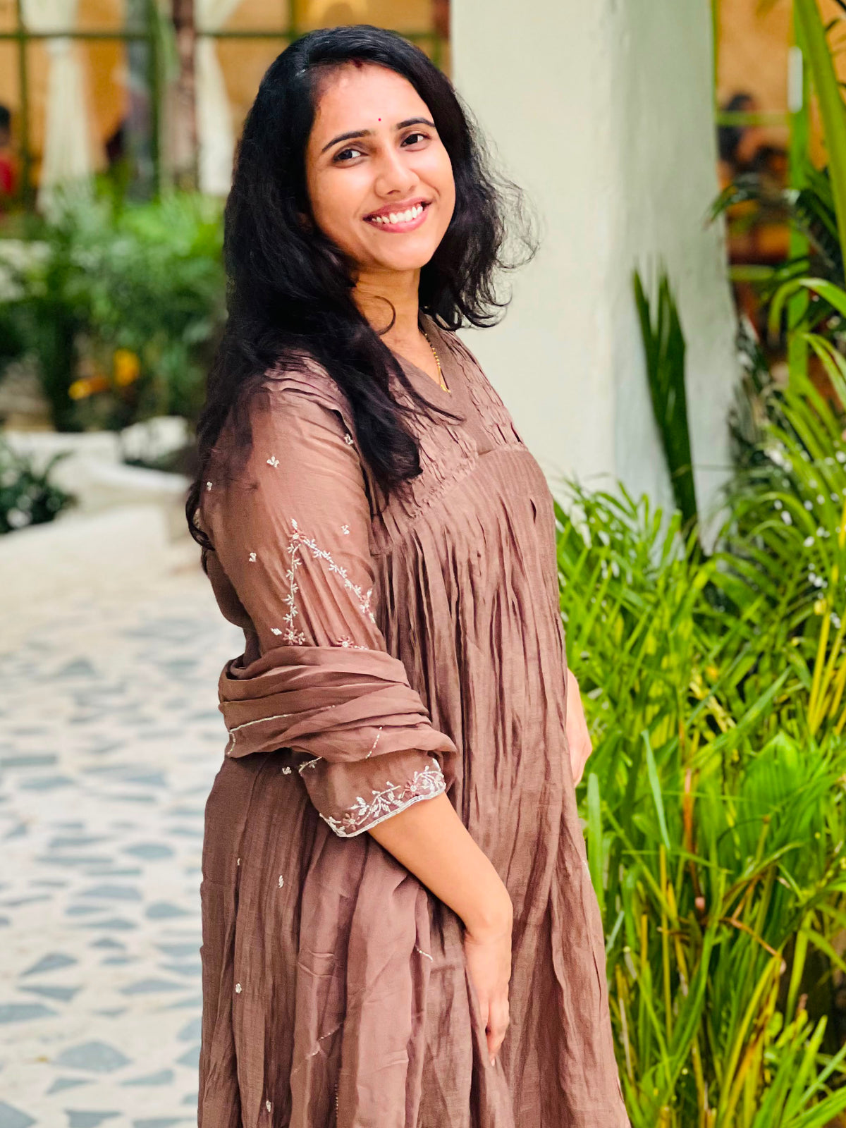A woman wearing a brown, semi-sheer, long-sleeved maxi dress with embroidered details on the sleeves, standing in a garden setting with lush green plants.